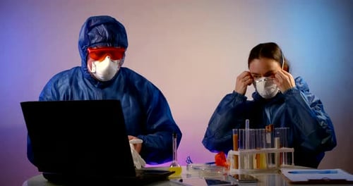 un hombre y una mujer con trajes de protección azules están sentados en una mesa con un ordenador portátil y tubos de ensayo.
Ellos