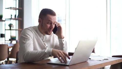 Busy Young Man Is Talking on Mobile Phone and Typing on Laptop in Office at Desk