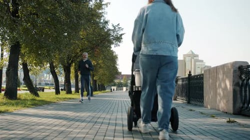 Young Mother Walking with Baby Carriage in City Center While Sportspeople are Running Around