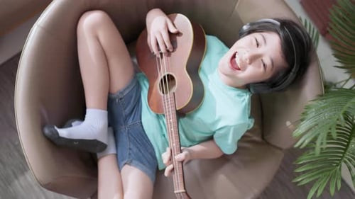 Smiling Child Plays Ukulele in Chair Indoors