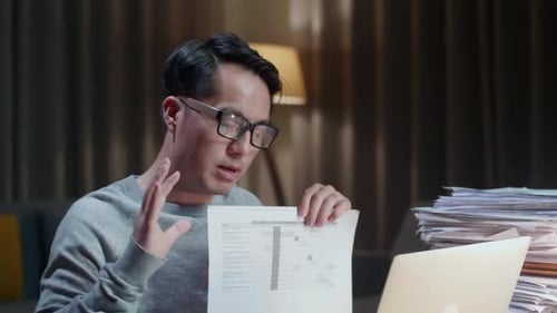 Close Up Of Asian Man With Documents Explaining Work On Paper While Having Video Call On Laptop