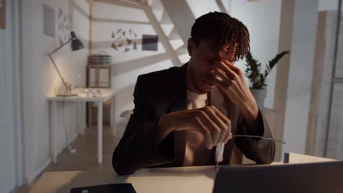 Overworked Afro-American Man Rubbing Nose and Using Laptop in Office