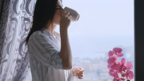 Woman Drinking Coffee by Apartment Window Overlooking City