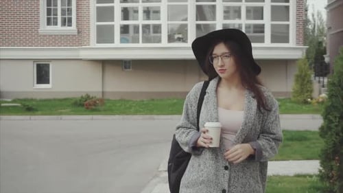 Pretty Woman in Black Hat and Glasses with Cup of Coffe Walking Down the Street