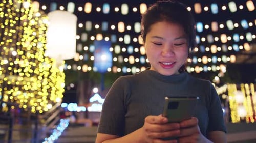Smiling Woman Using Smartphone Amidst Festive Lights