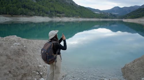 Girl with Hat and Backback Seen From Behind Films Lake with Phone