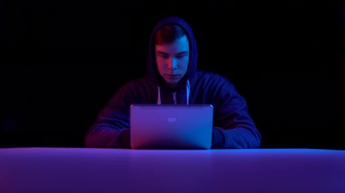 Young Man in a Hood with a Laptop. Hacker Makes a Hack Through a Laptop. Blue and Red Light Falls on