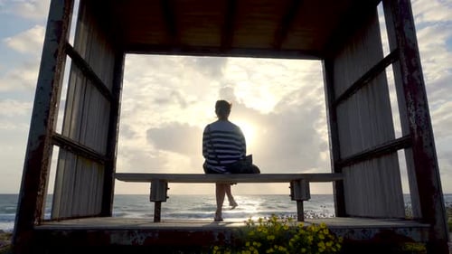 Woman Sits Silhouetted on Bench Overlooking Ocean at Sunset