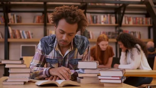 Dark Skinned Young Male Sitting at Table in Library Surrounded By Books Making Notes While Two