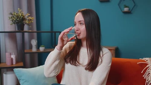 Portrait of Young Woman Sitting on Sofa at Home Holds Glass Drinks Water Makes Gulp of Clean Aqua
