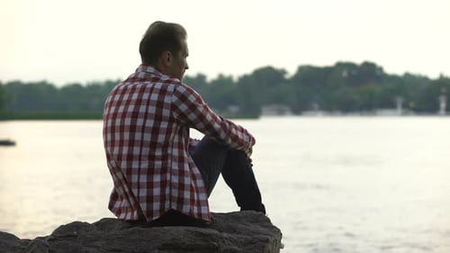 Depressiver erwachsener Mann sitzt am Flussufer und denkt über Scheidung nach, Einsamkeit