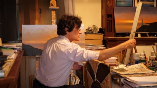 An artist paints an oil painting on canvas inside his art studio.