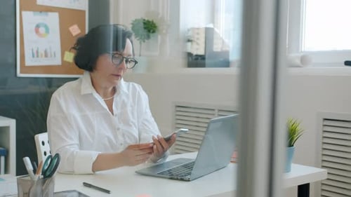 Cheerful Woman Using Laptop Typing Then Talking on Mobile Phone and Smiling Working in Office Alone