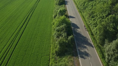Couple of Cyclists Rides on Asphalt Road Past Planted Field