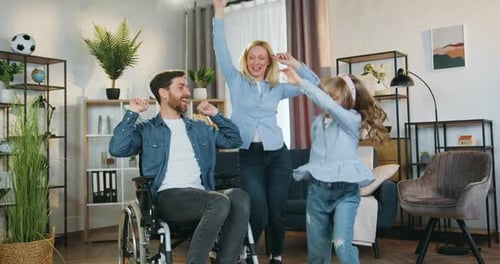 Hombre discapacitado en silla de ruedas viendo cómo bailan su alegre y encantadora esposa y su linda hija de 10 años