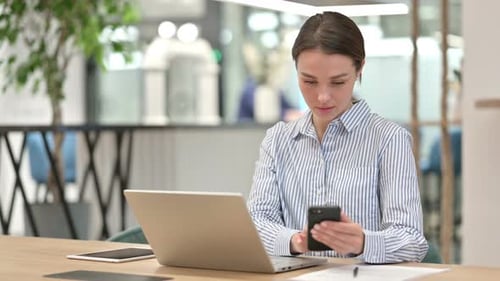 Young Woman with Laptop Using Smartphone in Office