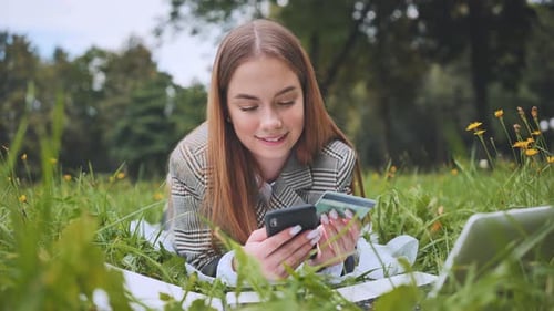 A Young Girl Makes a Purchase with Her Phone and Bank Card While Lying on the Grass in the Park