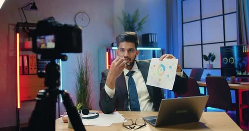 Empresario en traje sentado frente a la cámara en un trípode en la oficina nocturna y explicando el gráfico