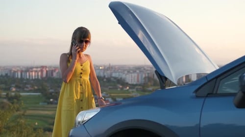 Stranded female driver talking on cell phone standing near her vehicle with open hood having