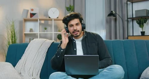 Hombre con auriculares sentado en el sofá de casa y disfrutando de su música favorita cantando canciones ante la cámara
