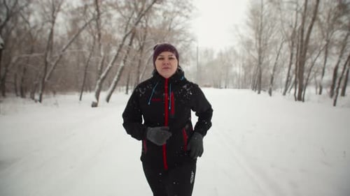 Caucasian Candid Woman in Black Jacket and Hat Runs Through Park in Winter Doing Daily Cardio