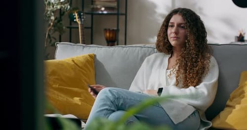 A Smiling Woman Sits on the Couch in Front of the TV Turns on a Movie on the Screen with the Remote
