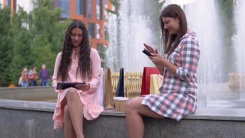 Two Young Girls After Shopping are Sitting Near the Fountain in the Park