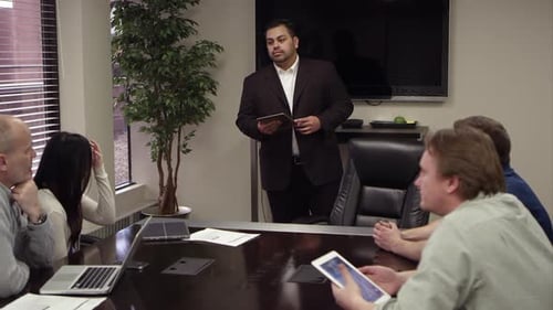 Panning view of people at conference table with guy standing and talking.