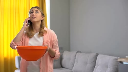 Upset Woman Calling Roof Repair Service While Water Leaking From Ceiling in Pot