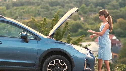 Puzzled young female driver standing near a broken car with open up hood inspecting her