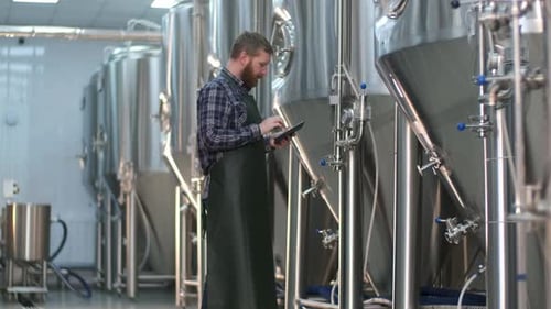Ein männlicher Brauer mit Bart überprüft die Messwerte von Geräten an Biertanks mit einem Tablet. Craft Bier