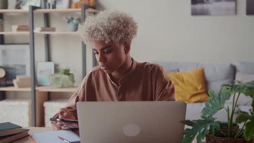 Young African American Woman Student Holding Phone Sits at Table with Laptop
