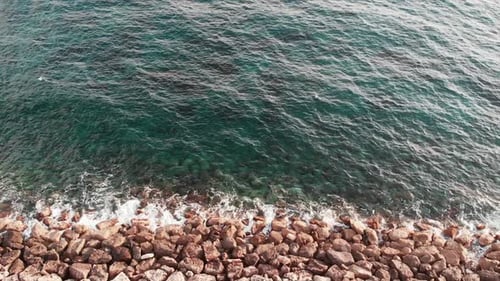 Aerial view of ocean waves hitting rocky beach creating foam. Clear blue sea collapse with rocks