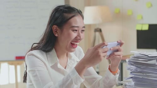 Primer plano de mujer asiática disfruta jugando en smartphone después de trabajar con documentos en la oficina