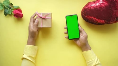 Hand Holding Smart Phone with Green Screen and Gift on Yellow Background