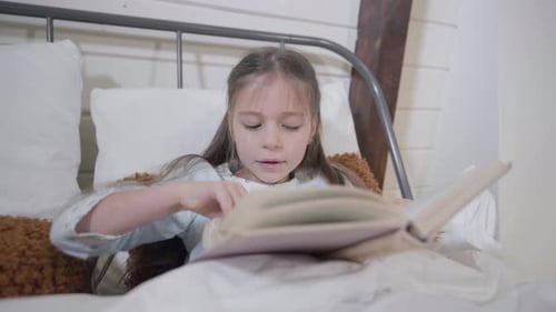 Portrait of Pretty Caucasian Girl Lying in Bed and Reading Book. Smart Child Going for Sleep in the