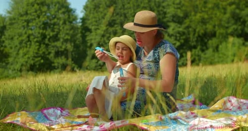 Funny Mom and Little Daughter Blow Soap Bubbles Sitting on the Lawn in the Park