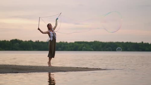 A Young Female Artist Shows a Soap Bubble Show Blowing Up Huge Soap Bubbles on the Shore of a Lake
