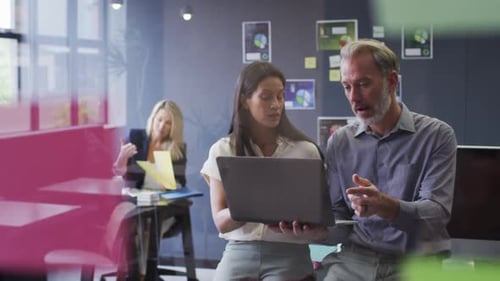 Two diverse business people using laptop talking in office and colleague working behind
