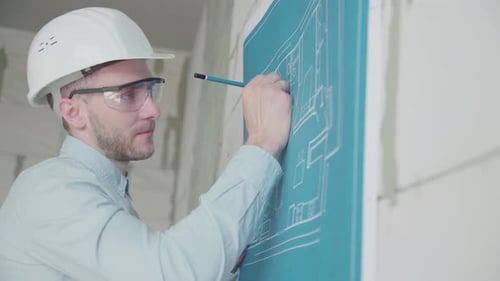 Young engineer-architect in a bright room in a helmet and glasses works with blueprints