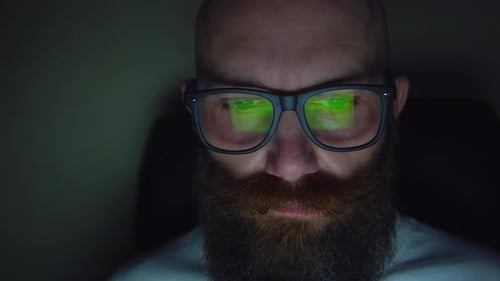 Homme barbu à lunettes naviguant sur le site Web tard dans la nuit, reflet à des lunettes