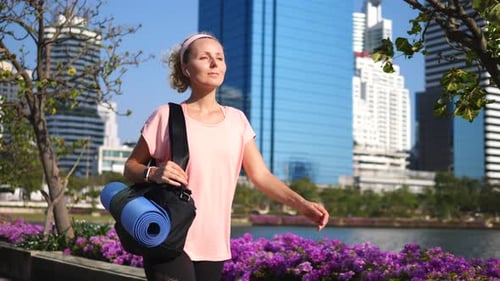 Fitness Woman Walking In City Park With Yoga Mat Wearing Wireless Earphones
