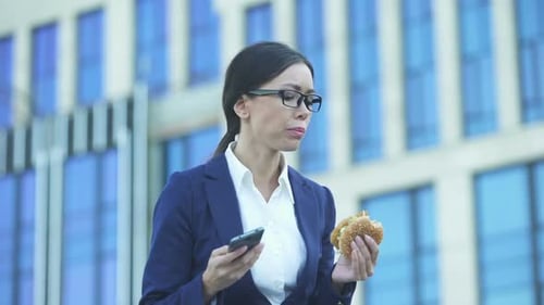 Female Office Manager Eating Unhealthy Burger Checking Messages on Phone, Stress