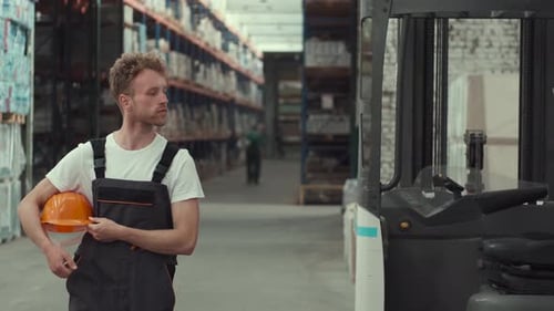 Worker with Helmet in Hands Standing at the Storehouse Near the Forklift Truck