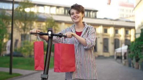 Mujer joven encantadora y segura de sí misma que camina con un scooter y bolsas de compras colgando