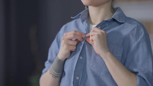 Irreconocible joven caucásica abotonándose camisa azul en interiores