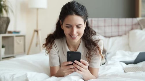 Woman Smiling While Using Phone on Bed