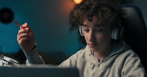A Boy with Curly Hair Sits in a Room at a Desk with a Laptop on Wireless Headphones Taking Notes