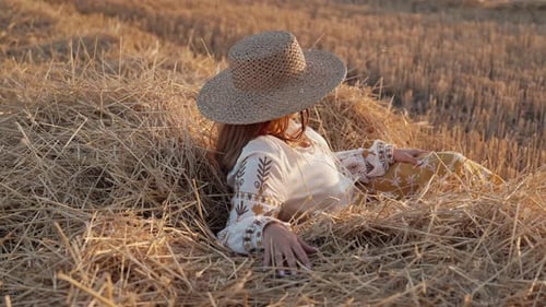 Beautiful Ukrainian Woman in Straw Hat and Vyshyvanka Blouse Posing Lying on Hay in Countryside at