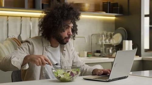 Young Adult Eating Salad While Working on Laptop